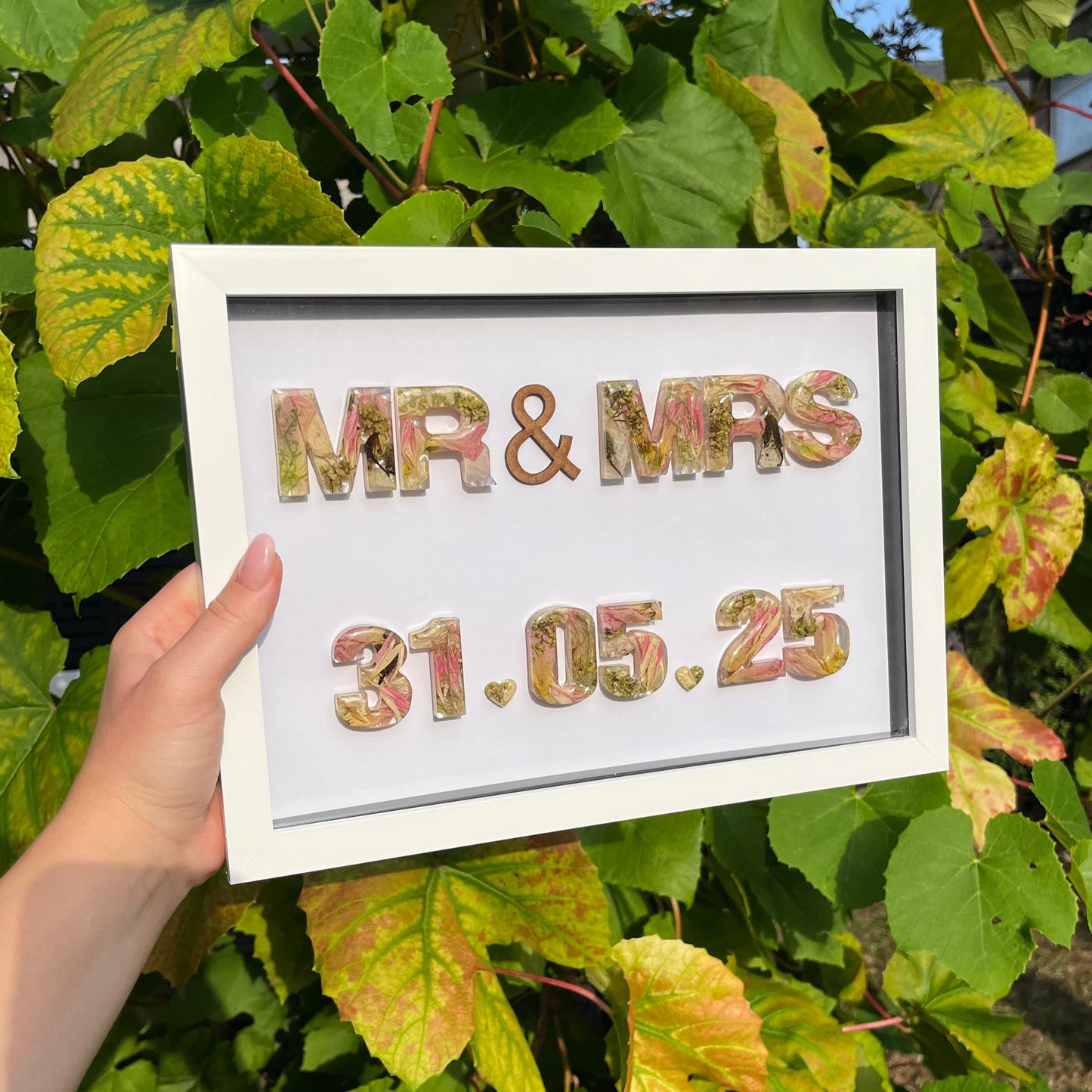 Framed decorative sign with 'MR & MRS' and a date, held in front of green foliage.