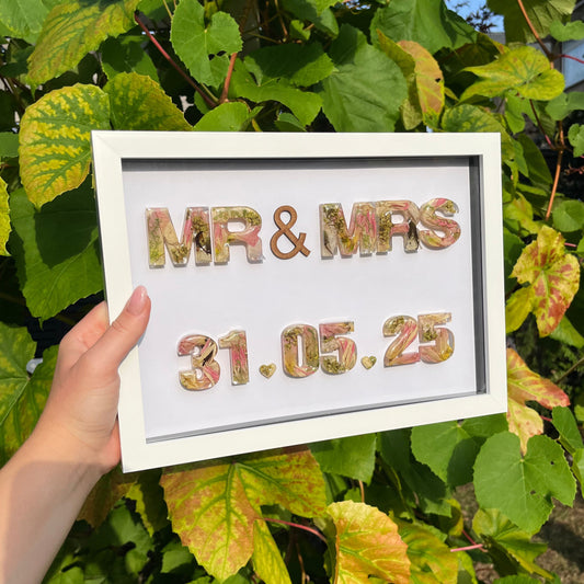 Framed decorative sign with 'MR & MRS' and a date, held in front of green foliage.