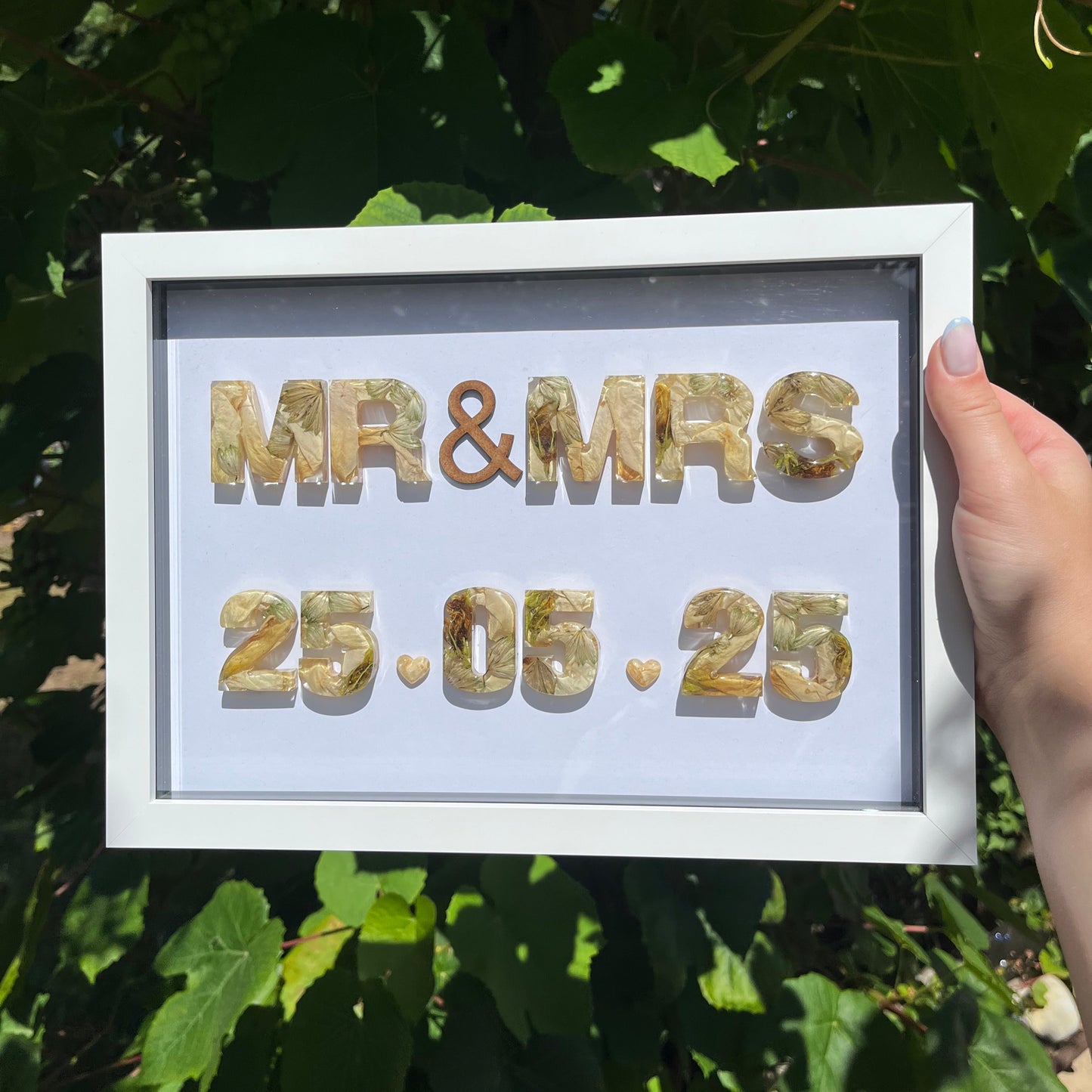 Framed resin letters spelling 'MR & MRS' and '25.06.25' against a green leafy background.