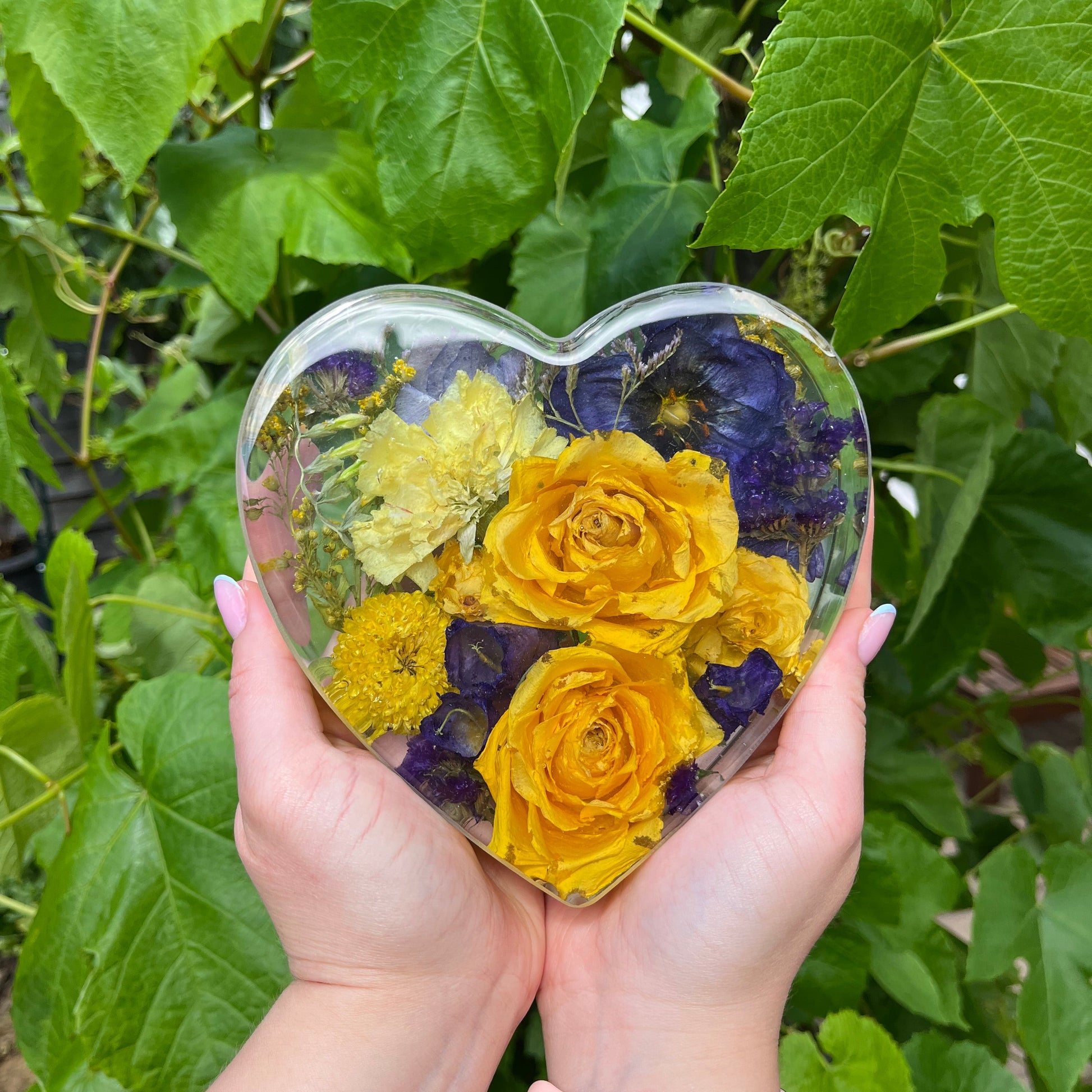 Heart-shaped clear block with yellow and purple flowers held by a person against a green leafy background