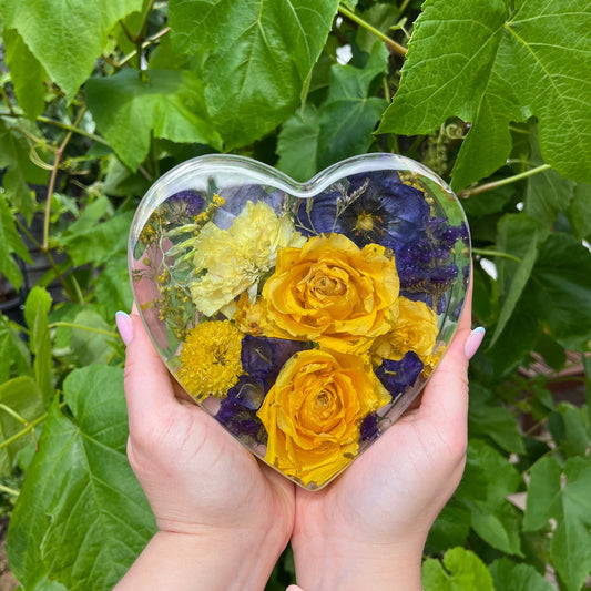 Heart-shaped clear block with yellow and purple flowers held by a person against a green leafy background