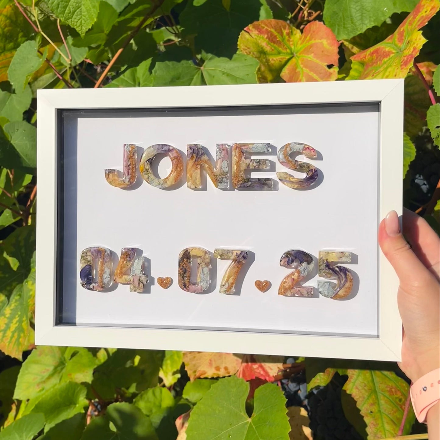 Framed artwork with 'JONES' and '04.07.25' text held in front of green leaves