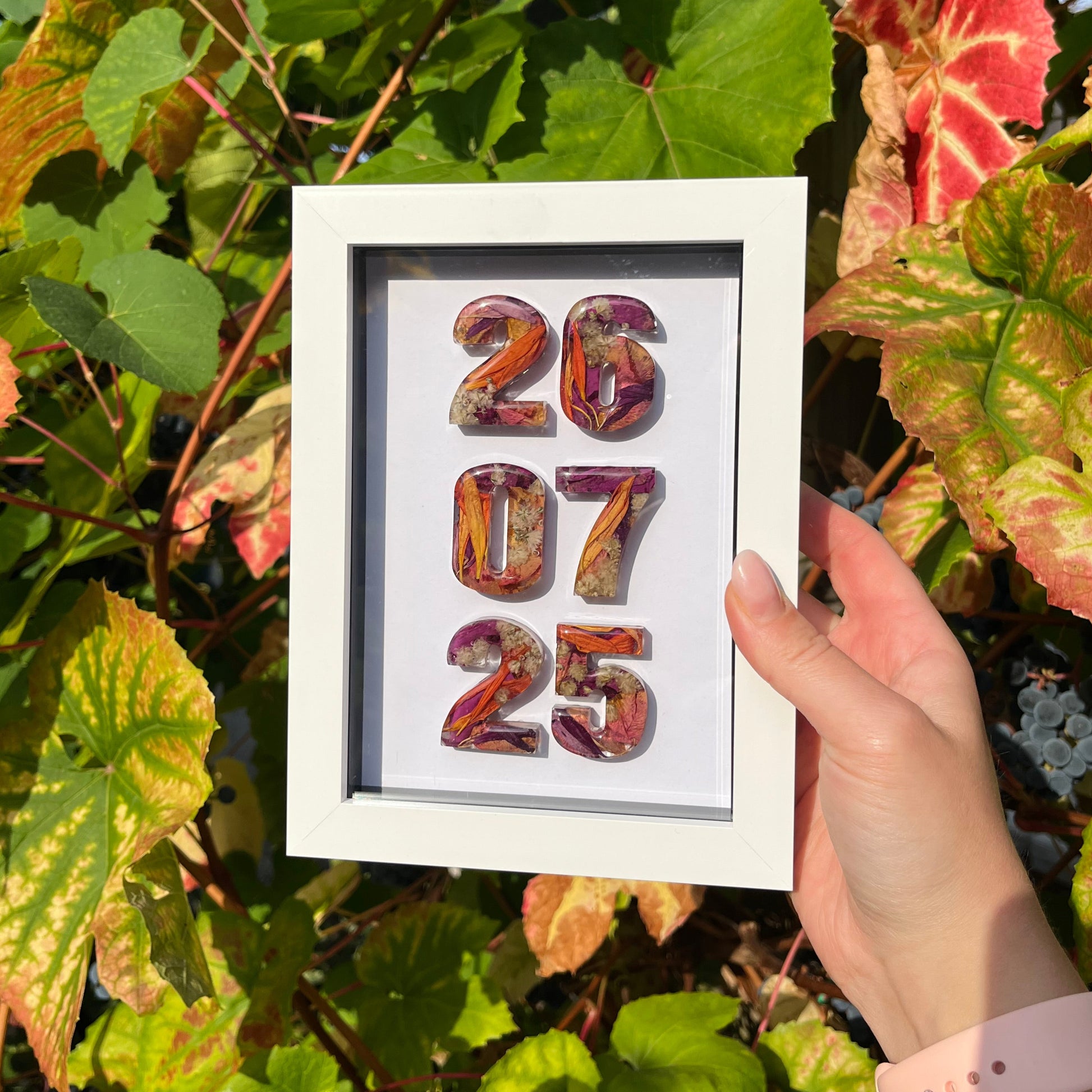 Framed artwork with colorful numbers held by a hand among green leaves