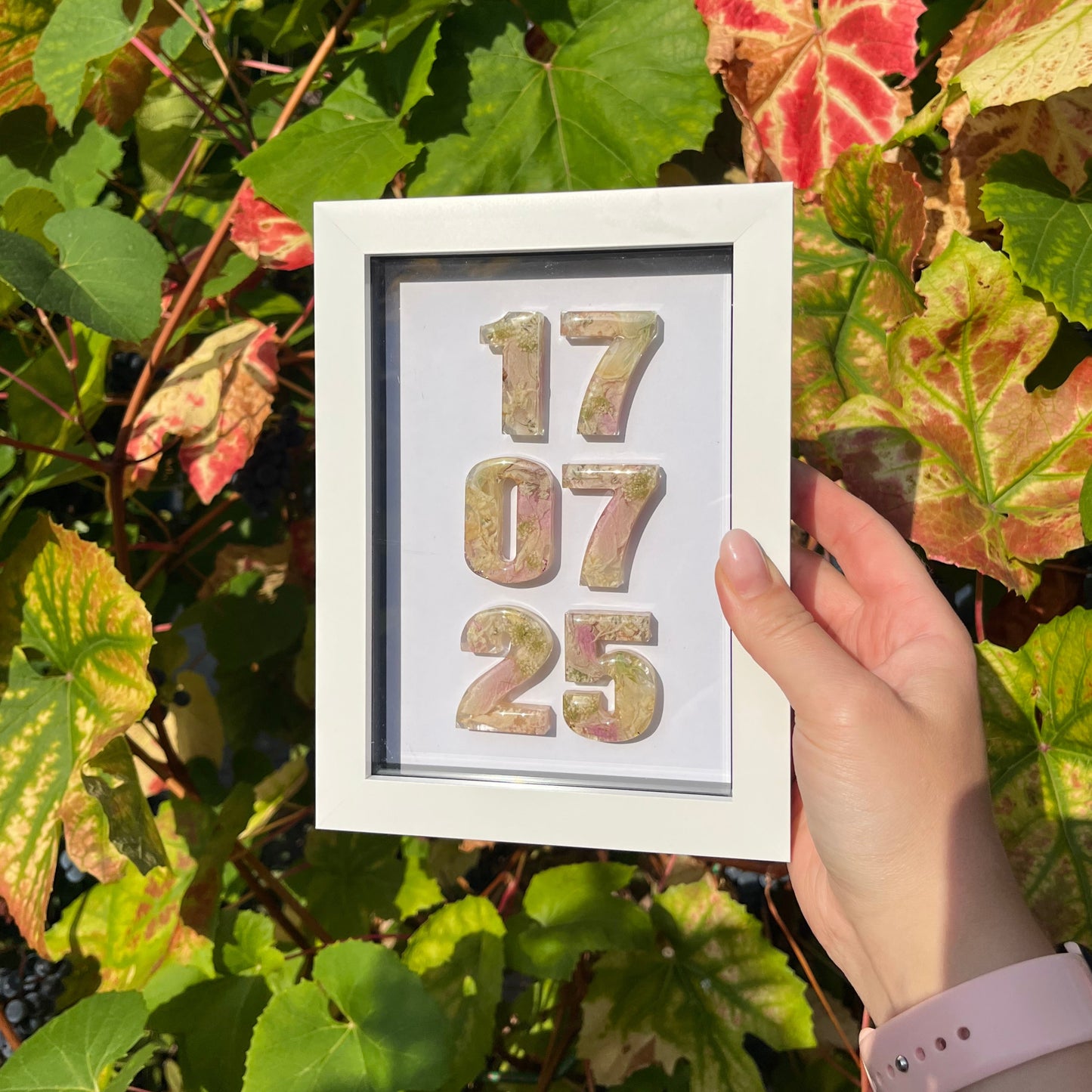 Framed numbers '17.07.25' held by a hand among green leaves