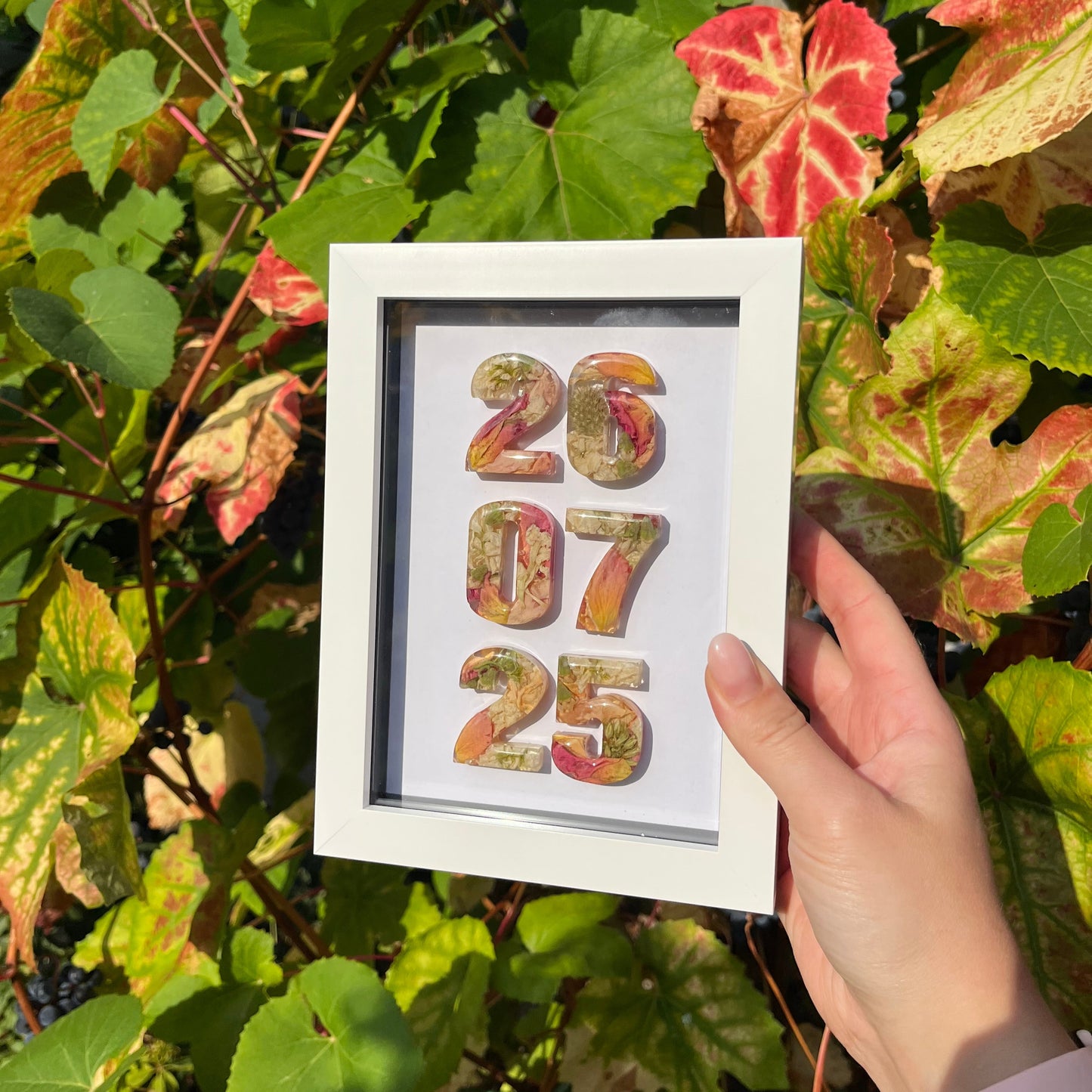 Framed artwork with colorful numbers held by a hand among green leaves