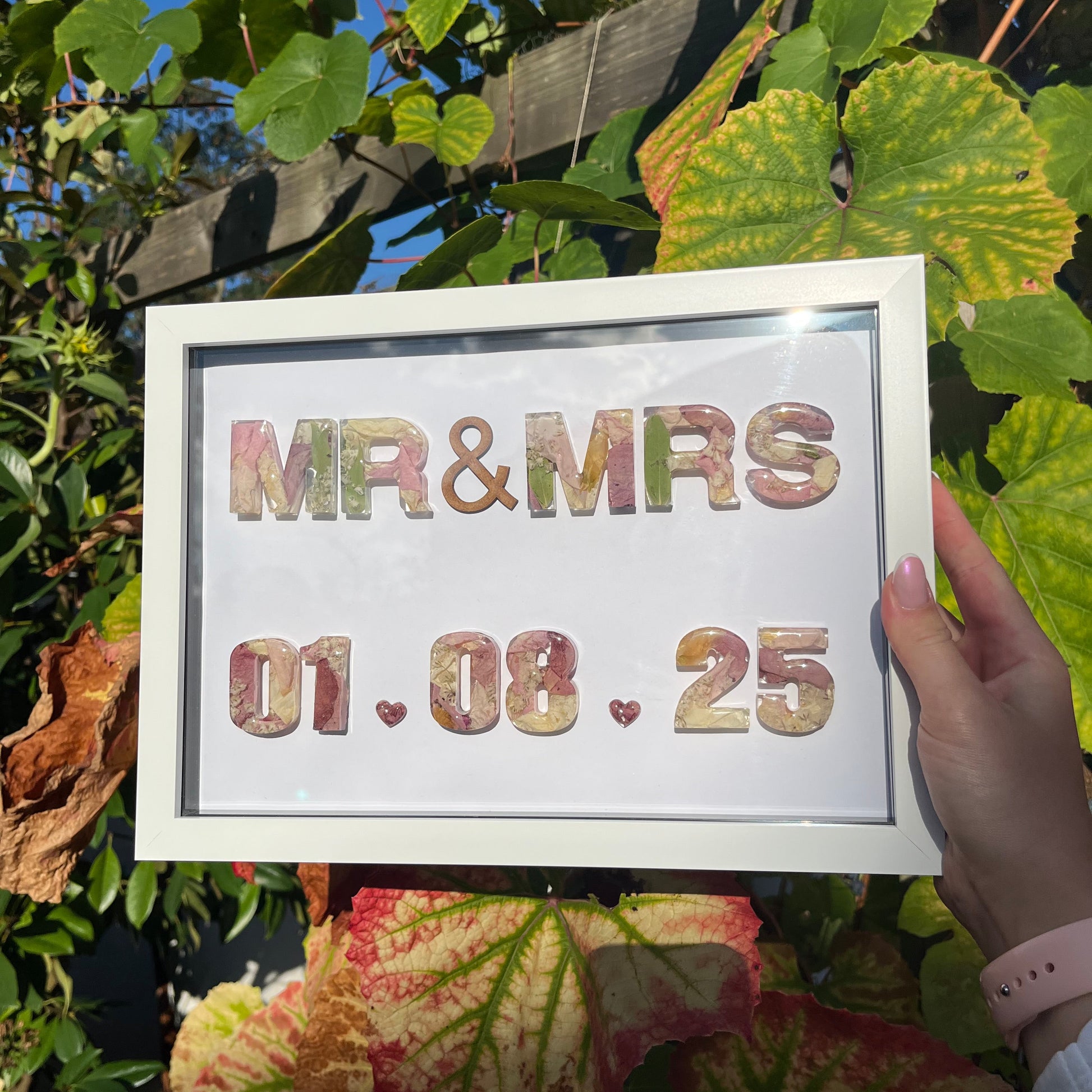 Framed sign with 'MR & MRS' and a date, held by a hand among green leaves.