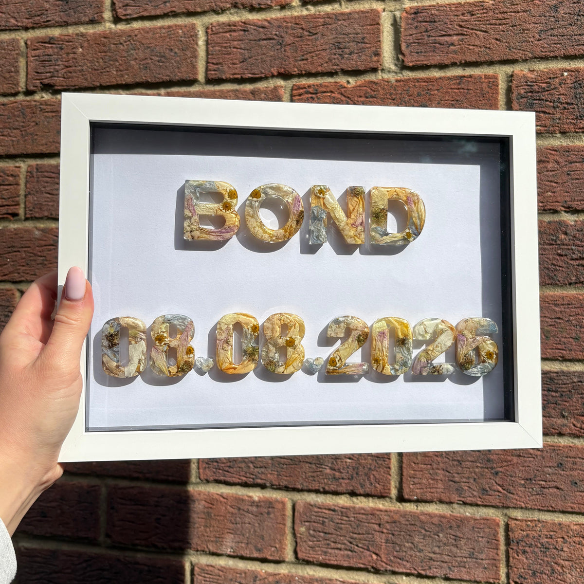 A hand holds a white shadow box frame against a brick wall. The frame contains custom floral lettering spelling 'BOND' and the date '08.08.2026'