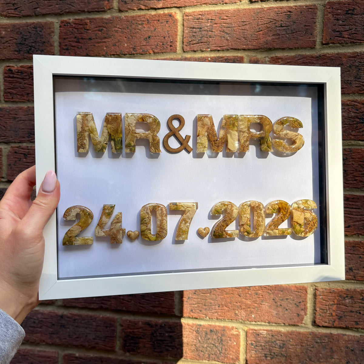 A hand holds a white framed custom shadow box featuring the words 'MR & MRS' and the date '24.07.2026'