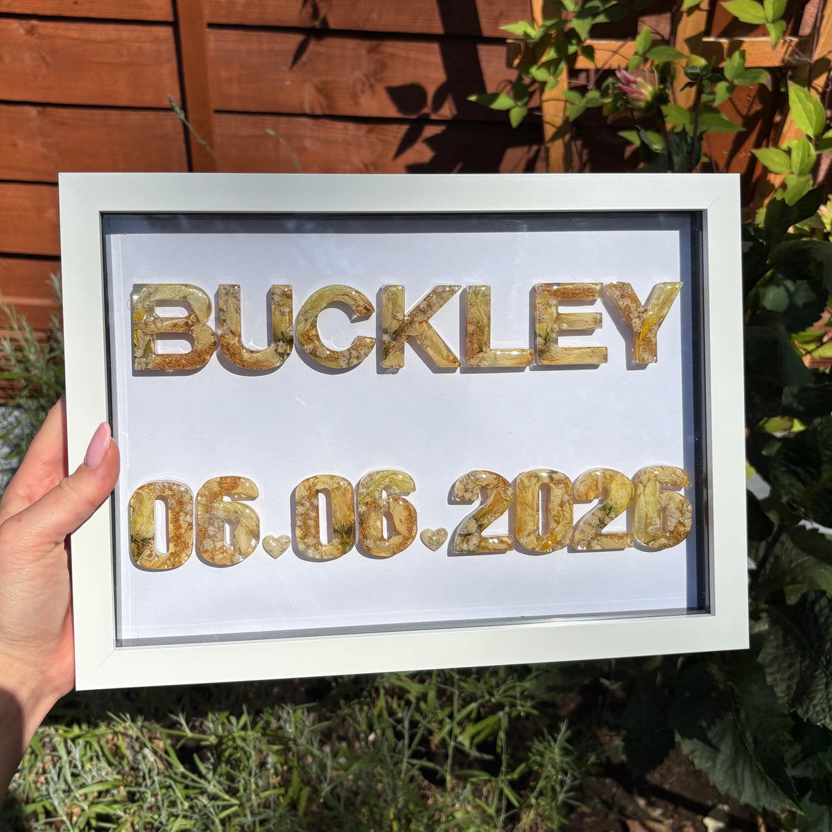 A hand holds a white shadow box frame outdoors in front of a wooden fence and green plants. The frame contains the name 'BUCKLEY' in floral resin letters and the date '06.06.2026'