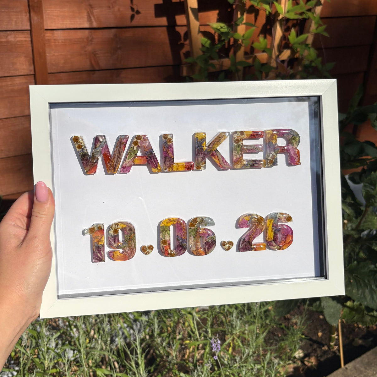 A hand holds a white-framed floral resin letters featuring the name 'WALKER' and the date '19.06.26'