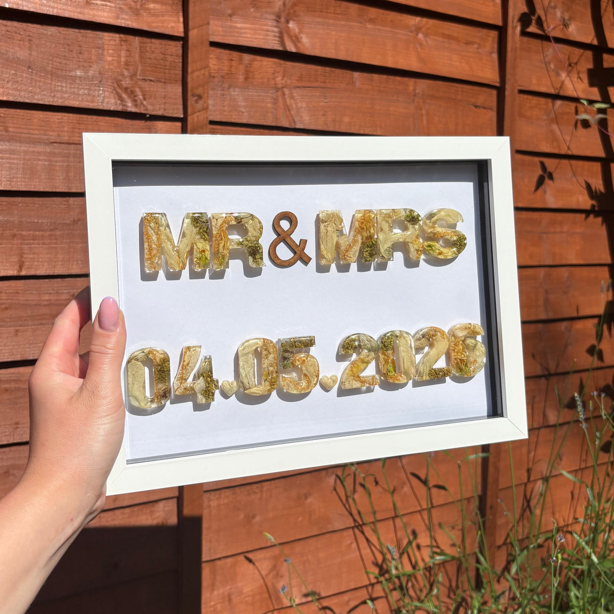 A hand holds a white framed custom sign with floral letters spelling 'MR & MRS' and the date '04.05.2026'
