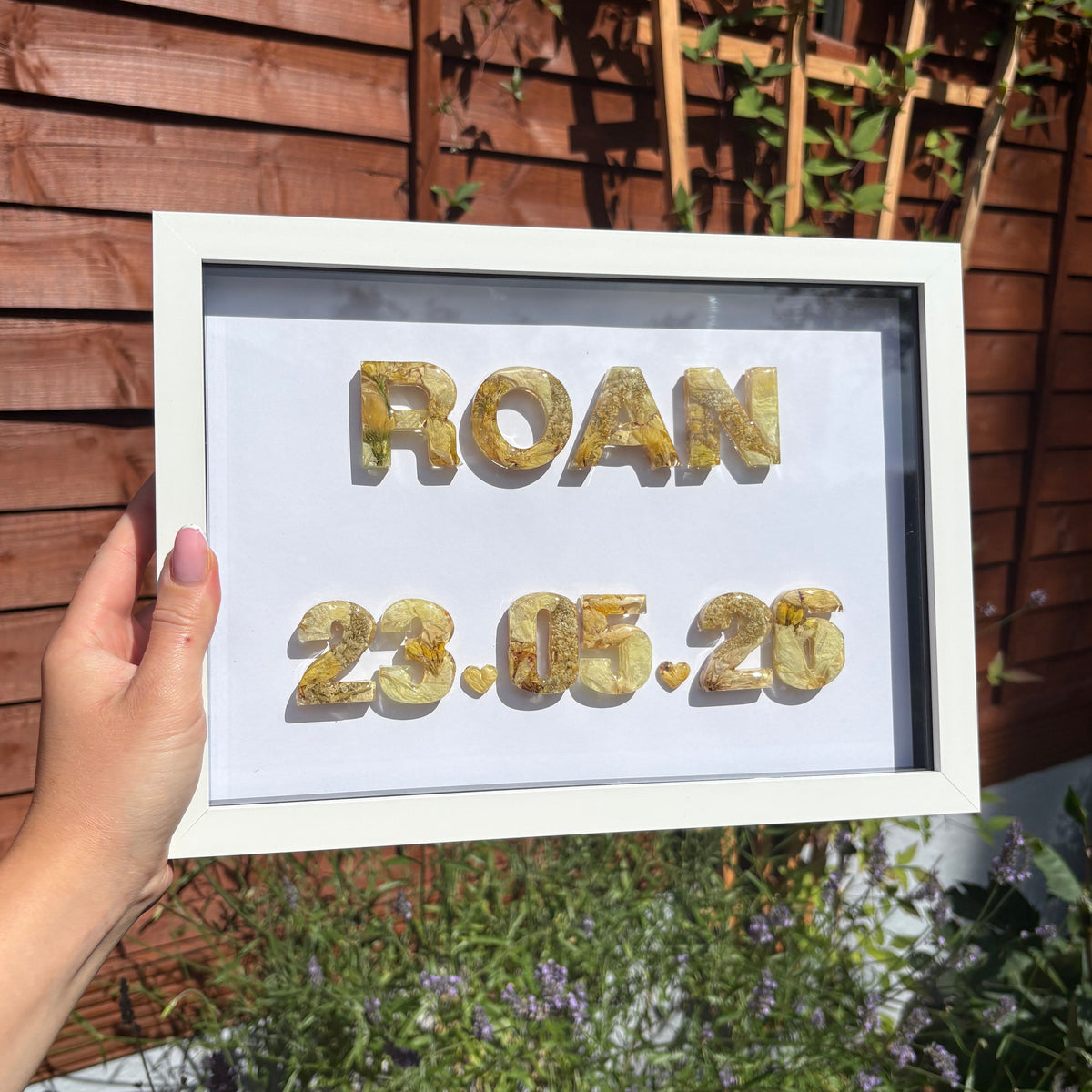 A hand holds a white-framed in a garden setting. The white background of the frame features floral letters spelling 'ROAN' and the date '23.05.26'